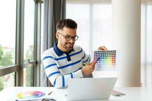 vrolijk Mens ontwerper hebben video conferentie met klanten, zittend Bij bureau in voorkant van computer, Holding kleur paletten, gebaren en lachend, kopiëren ruimte foto