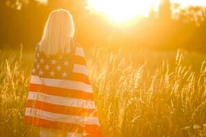 mooi jong meisje Holding een Amerikaans vlag in de wind in een veld- van rogge. zomer landschap tegen de blauw lucht. horizontaal oriëntatie. foto