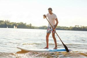 portret van een surfer met een sup bord Aan de strand. jong Mens Aan paddleboard Bij ochtendgloren. de concept van extreem sport. mannetje surfer levensstijl. foto