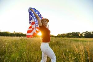 mooi jong meisje Holding een Amerikaans vlag in de wind in een veld- van rogge. zomer landschap tegen de blauw lucht. horizontaal oriëntatie. foto