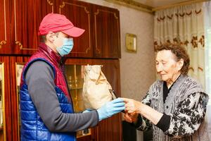 jong mannetje vrijwilliger in masker geeft een ouderen vrouw dozen met voedsel in de buurt haar huis. zoon Mens helpt een single ouderen moeder. familie steun, zorgzaam. in quarantaine geplaatst, geïsoleerd. coronavirus covid19. bijdrage foto