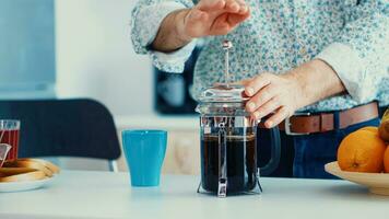 oud Mens voortvarend naar beneden de deksel van Frans druk op terwijl maken koffie voor ontbijt in keuken. ouderen persoon in de ochtend- genieten van vers bruin cafe espresso kop cafeïne van wijnoogst mok, filter kom tot rust verfrissing foto