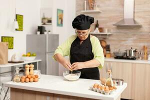 ouderen gebakje chef kraken ei Aan glas kom voor taart recept in keuken., menging door hand, kneden ingrediënten voorbereiden eigengemaakt taart foto