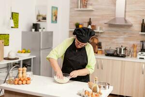 vrouw menging eieren met meel naar maken deeg na traditioneel recept Aan keuken tafel. gepensioneerd ouderen chef met uniform besprenkelen, zeven zeven rauw ingrediënten en menging . foto