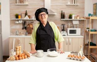 oud dame vervelend chef schort en bot in huis keuken met meel Aan tafel. gepensioneerd ouderen bakker in keuken uniform voorbereidingen treffen gebakje ingrediënten Aan houten tafel klaar naar koken eigengemaakt smakelijk brood. foto