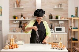 senior dame chef verspreiden meel met hand- voor voedsel voorbereiding in huis keuken vervelend schort. gelukkig ouderen chef met uniform besprenkelen, zeven zeven rauw ingrediënten door hand- bakken eigengemaakt pizza. foto