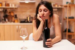 vrouw zittend alleen met een fles van rood wijn in keuken omdat van onderdrukking. ongelukkig persoon lijden van migraine, depressie, ziekte en ongerustheid gevoel uitgeput. foto