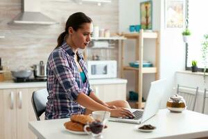 vrouw werken van huis gebruik makend van laptop in keuken gedurende ontbijt. werken van huis gebruik makend van apparaat met internet technologie, browsen, zoeken Aan apparaatje in de ochtend. foto