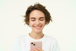 dichtbij omhoog portret van jong vrouw lachend, gebruik makend van smartphone, sms'en, controle haar mobiel appjes, staat over- wit achtergrond foto