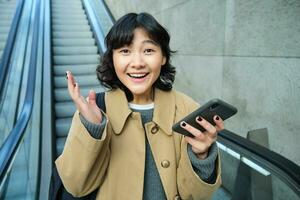 gelukkig zegevieren Aziatisch meisje, looks versteld staan en blij, houdt smartphone, gaat naar beneden roltrap met verheugd gezicht, wint, hoort mooi zo nieuws foto