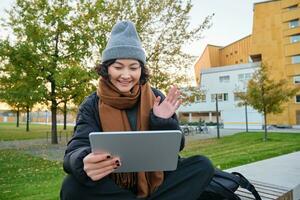 portret van jong Aziatisch meisje zit in warm kleren in park, golven hand- Bij tablet, video chats buitenshuis, zegt Hallo terwijl Aan telefoontje foto