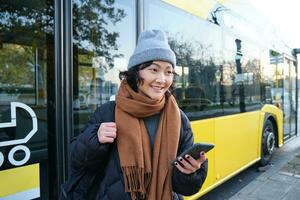 portret van meisje staand in de buurt bus Aan een hou op, aan het wachten voor haar openbaar vervoer, cheques schema Aan smartphone sollicitatie, houdt mobiel telefoon, draagt warm kleren foto