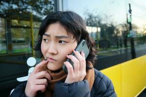 perplex Aziatisch meisje, praat Aan mobiel telefoon met bezorgd, attent gezicht, staat in de buurt bus hou op en fronst, denken terwijl luisteren telefoon gesprek foto