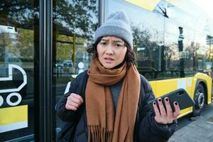 portret van verward Aziatisch meisje, staand Aan bus hou op, Holding mobiel telefoon, op zoek geschokt en van streek, teleurgesteld door openbaar vervoer vertraging foto