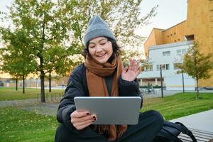 portret van jong Aziatisch meisje zit in warm kleren in park, golven hand- Bij tablet, video chats buitenshuis, zegt Hallo terwijl Aan telefoontje foto