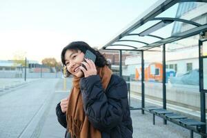 mooi glimlachen Koreaans meisje, aan het wachten Aan bus hou op, gebruik makend van openbaar vervoer, pratend Aan mobiel telefoon, gaan ergens in stad foto