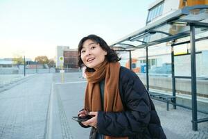 schattig jong meisje, leerling aan het wachten Aan een bus hou op, op zoek voor haar openbaar vervoer Aan app volger, Holding smartphone, staand Aan straat in de buurt weg foto