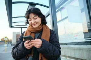 mooi Japans meisje, leerling staand Aan bus hou op in winter jasje, Holding smartphone, gebruik makend van toepassing naar bijhouden haar openbaar vervoer, bestellen een rijden foto