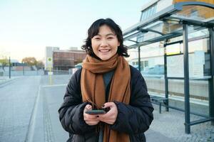 portret van Koreaans vrouw in winter jasje, staand met smartphone, aan het wachten voor bus Aan hou op, op zoek Bij mobiel telefoon app controle openbaar vervoer toepassing foto