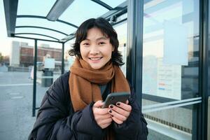 schattig jong meisje, leerling aan het wachten Aan een bus hou op, op zoek voor haar openbaar vervoer Aan app volger, Holding smartphone, staand Aan straat in de buurt weg foto