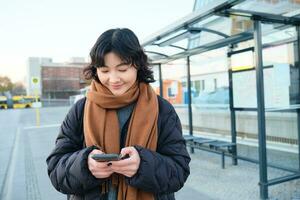 gelukkig glimlachen Koreaans meisje, gebruik makend van mobiel telefoon, staand Aan bus hou op met smartphone, op zoek Bij vertrek schema Aan sollicitatie, poseren in winter kleren foto