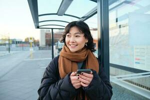 portret van Koreaans vrouw in winter jasje, staand met smartphone, aan het wachten voor bus Aan hou op, op zoek Bij mobiel telefoon app controle openbaar vervoer toepassing foto