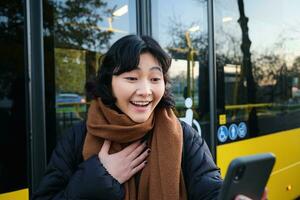 portret van vrolijk Aziatisch meisje praat Aan mobiel telefoon, video chatten, looks versteld staan Bij smartphone camera, staat Aan bus hou op foto