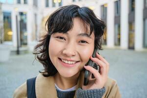 portret van glimlachen Aziatisch meisje praat Aan mobiel telefoon, spreekt met vriend Aan smartphone, wandelingen in stad centrum, staat Aan straat en lacht foto