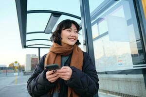 schattig glimlachen Aziatisch meisje staand Aan bus hou op, Holding smartphone, vervelend winter jasje en sjaal. vrouw woon-werkverkeer naar werk of Universiteit via openbaar vervoer, staat Aan weg foto