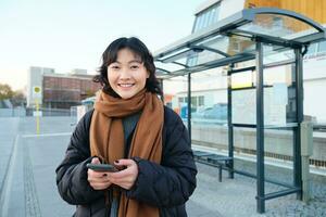 gelukkig glimlachen Koreaans meisje, gebruik makend van mobiel telefoon, staand Aan bus hou op met smartphone, op zoek Bij vertrek schema Aan sollicitatie, poseren in winter kleren foto