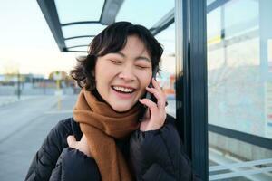 dichtbij omhoog van Aziatisch meisje Aan bus hou op, lacht over- telefoon gesprek, praat Aan telefoon terwijl wacht voor openbaar vervoer, draagt winter kleren foto