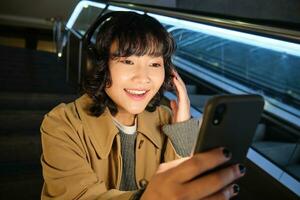 portret van gelukkig Aziatisch meisje in koptelefoon, glimlachen en op zoek Bij smartphone, aan het kijken video of luisteren muziek- Aan mobiel telefoon, zittend Aan trappenhuis foto