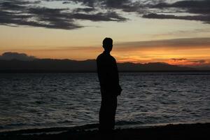 silhouet van een jong Mens staand door de meer genieten van de zonsondergang. vredig atmosfeer in natuur foto