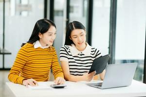 twee aantrekkelijk jong Aziatisch vrouw college studenten werken Aan de school- project gebruik makend van laptop computer en tablet samen, genieten pratend en snacken foto