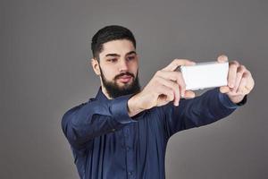 jonge, bebaarde hipster zakenman praten selfie foto met slimme telefoon glimlachen en kijken naar telefoon tegen grijze studio achtergrond
