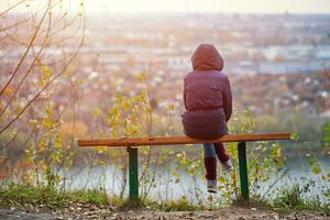 jong vrouw zittend Aan bank in herfst stad park en op zoek Bij stadsgezicht, terug visie foto