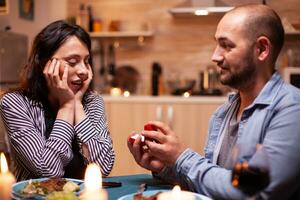 Mens vieren verhouding en vragen vriendin naar trouwen hem terwijl hebben diner. Mens vragen zijn vriendin naar trouwen in de keuken gedurende romantisch diner. gelukkig Kaukasisch vrouw glimlachen wezen sprakeloos foto