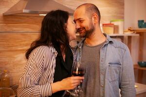 paar aanraken neuzen en hebben romance datum in keuken. volwassen paar Bij huis, drinken rood wijn, praten, lachend, genieten van de maaltijd in dining kamer. foto