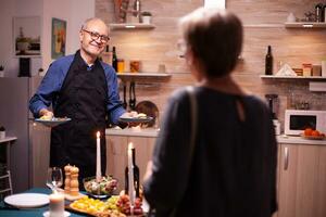 vrolijk senior Mens pratend met vrouw terwijl portie avondeten voor romantisch viering in keuken. ouderen oud paar praten, zittend Bij de tafel in keuken, genieten van de maaltijd. foto