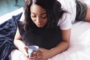mooi Afrikaans meisje in nachtkleding glimlachend kijkend naar camera met beker thuis zittend in bed
