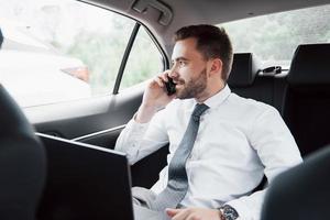 een jonge zakenman die aan een laptop werkt en aan de telefoon praat terwijl hij in de auto zit. werkt in beweging, waardeert zijn tijd