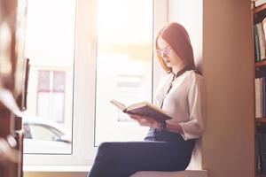 een studentenmeisje zit op een vensterbank in een bibliotheek en leest een boek foto