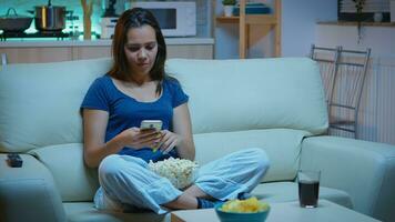 vrouw scrollen Aan telefoon aan het eten popcorn en aan het kijken een film. eenzaam geamuseerd gelukkig dame lezing, schrijven, zoeken, browsen Aan smartphone lachend grappig gebruik makend van technologie internet ontspannende Bij nacht. foto
