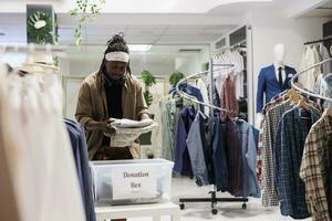 Afrikaanse Amerikaans Mens zetten kleding in bijdrage doos in boodschappen doen winkelcentrum. mode winkel klant inpakken kleding in houder voor liefdadigheid organisatie, geven kledingstuk voor arm foto