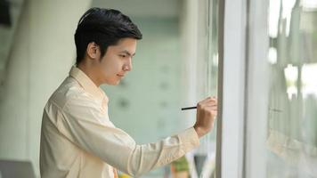 jonge zakenman maakt aantekeningen en werkt aan een notitie op een glazen wand in een modern kantoor.