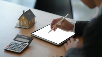 close-up shot van zakenmensen die een pen gebruiken om op een tablet te schrijven met een modelhuis en een rekenmachine op tafel.