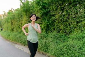 fit Aziatisch jong vrouw jogging in park glimlachen gelukkig rennen en genieten van een gezond buitenshuis levensstijl. vrouw jogger. geschiktheid loper meisje in openbaar park. gezond levensstijl en welzijn wezen concept foto