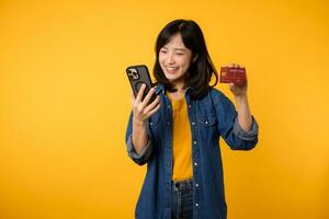 portret mooi jong Aziatisch vrouw gelukkig glimlach gekleed in denim jasje tonen smartphone en credit kaart isoleren Aan geel studio achtergrond. boodschappen doen online smartphone toepassing concept foto
