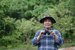 Aziatisch vrouw ontdekkingsreiziger draagt hoed, blauw plaid overhemd , houdt kijker naar ontdekken, enquête botanisch planten en schepsels dieren in het wild. concept, natuur verkenning. ecologie en omgeving. foto