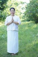 Aziatisch vrouw boeddhistisch draagt wit kostuum, zetten palmen samen voor bidden, staat Bij rustig Woud. vol lichaam afbeelding. concept, cultuur van vertrouwen. religieus werkzaamheid. vrede van geest beheer. foto